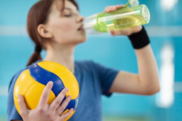 女子バレーボール選手がバレーボールコートでボトルから水を飲む バレーボールのトレーニング中に水を飲む女の子が手にボールに焦点を当てる