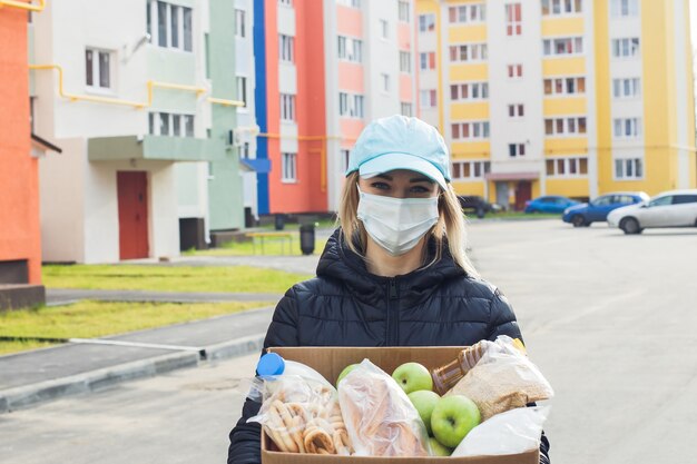 Женщина-волонтер в защитной маске идет по улице с коробкой продуктов, пожертвованиями.