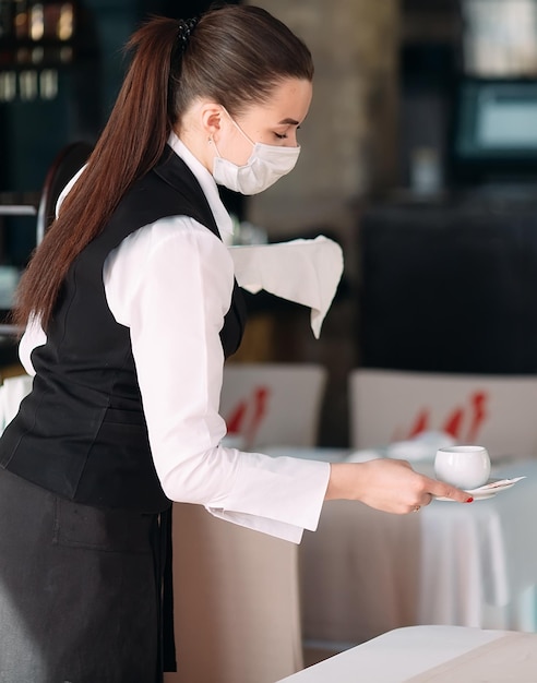 A female Waiter of European appearance in a medical mask serves Latte coffee.