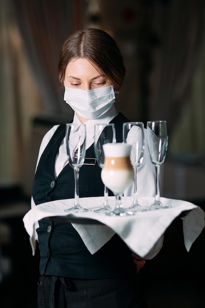 의료용 마스크를 쓴 유럽풍 여성 웨이터가 라떼 커피를 제공합니다.