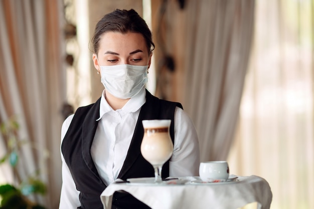 A female Waiter of European appearance in a medical mask serves Latte coffee.