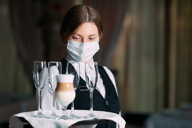 의료 마스크에 유럽 모양의 여성 웨이터가 라떼 커피를 제공합니다.