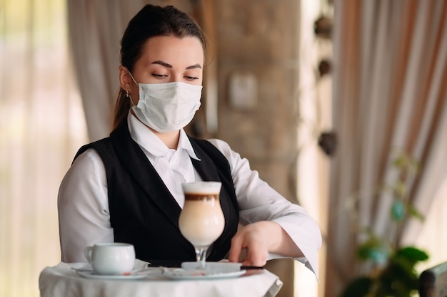 A female Waiter of European appearance in a medical mask serves Latte coffee.