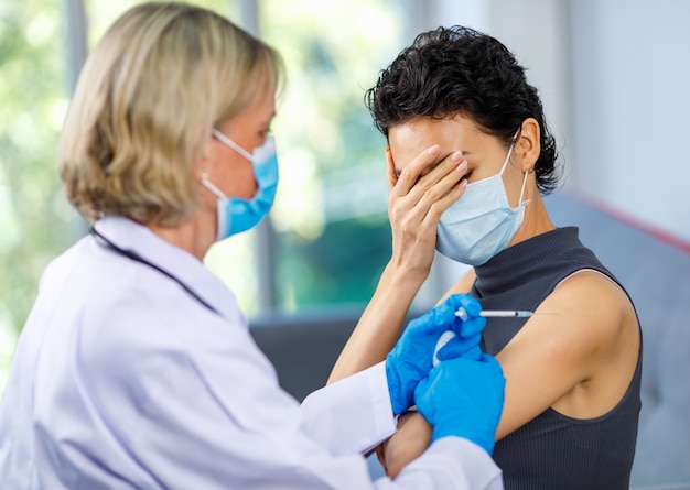 Female wears face mask sit hold hand close eye afraid of pain from vaccination while doctor using syringe injecting coronavirus vaccine. Vaccinophobia, needle phobia or Trypanophobia effect concept.