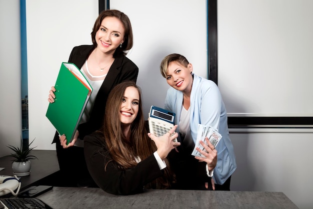 Female with long hair holding bundle dollars with calculator\
counting revenue three businesswoman at a work laughing while\
working at an office table womans with cash money in dollar\
banknotes