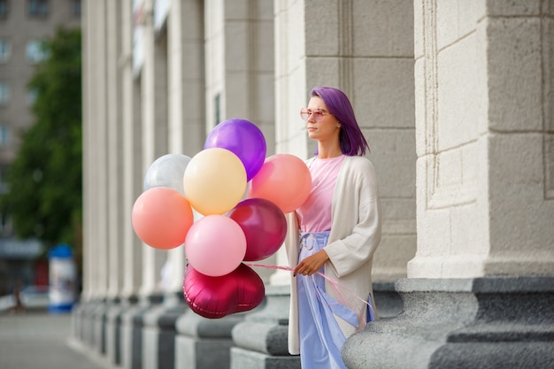 baloons의 무리와 함께 서있는 분홍색 안경에 보라색 머리를 가진 여성