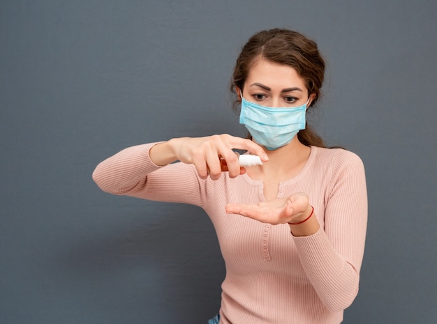 Female woman in medical mask on her face using sanitizer gel for disinfecting her hands on gray surface