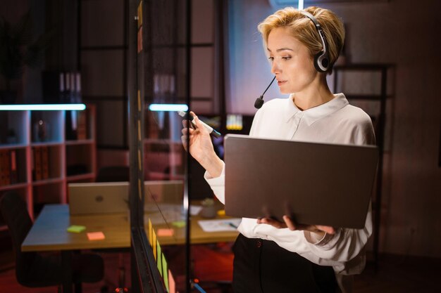 Photo female worker wearing headset and using laptop writing ideas and project tasks on glass wall