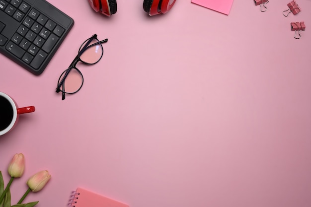 Female workspace with glasses, coffee cup, tulips and headphone on pink background.