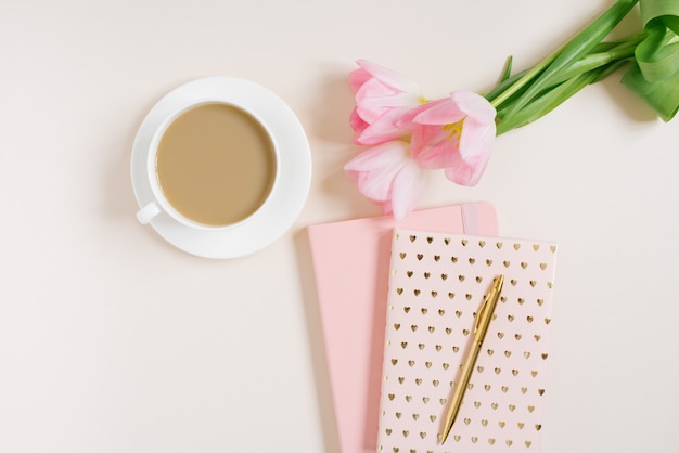 Female workspace with pink tulip flowers, diary, cup of tea and donut on beige background. Flat lay. Top view background.