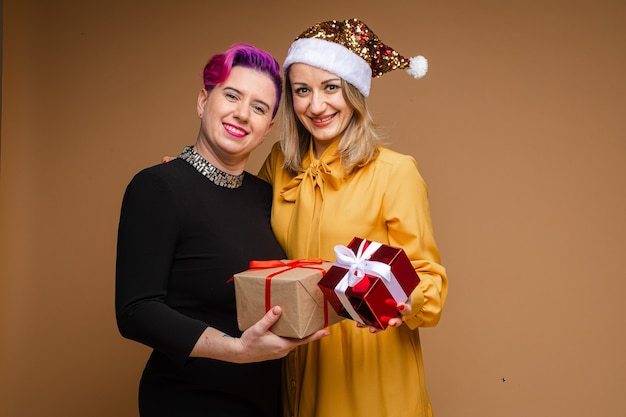 Female in yellow dress wrapping her arm around her girlfriend shoulder with them both smiling and showing their gifts. New Year concept