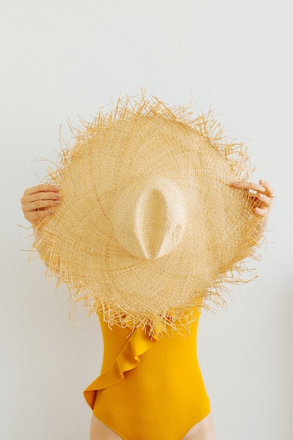 Female in a yellow swimsuit holds summer straw hat on white background