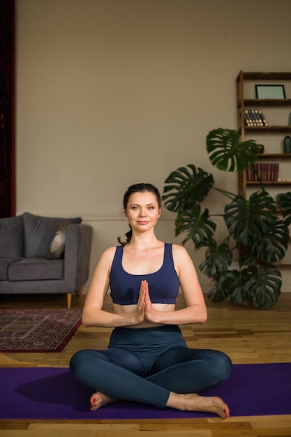 Female yogi in sportswear sits in a lotus position on a yoga mat in a room