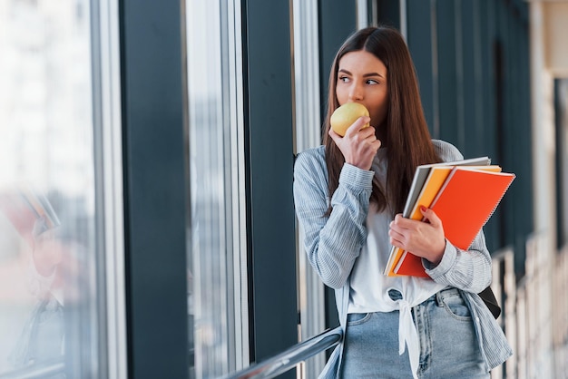 Female young student is in corridor of a college holding notepads and an apple