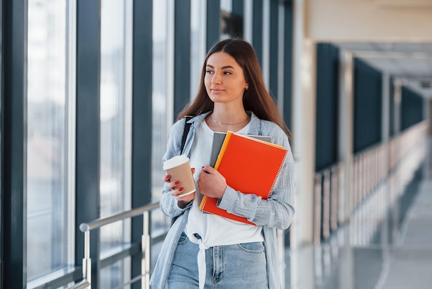 Female young student is in corridor of a college holding notepads and cup of drink