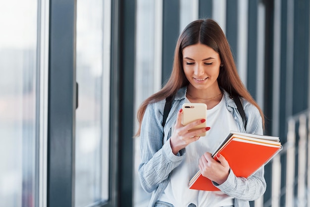 Female young student is in corridor of a college holding notepads and phone