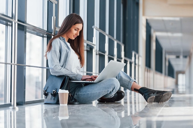 Female young student sitting in corridor of a college with laptop and cup of drink on floor