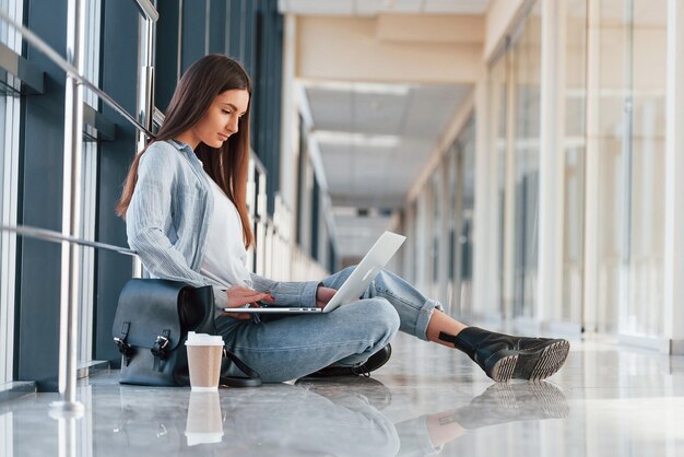 Giovane studentessa seduta nel corridoio di un college con laptop e tazza di bevanda sul pavimento