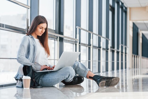 Female young student sitting in corridor of a college with laptop and cup of drink on floor