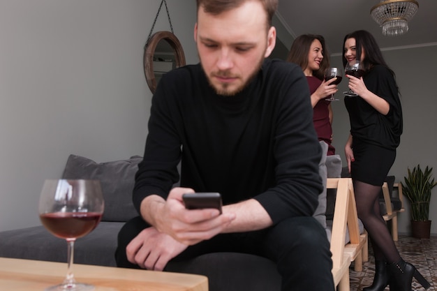 Females gossips about man at party. Funny women conversation in focus on background. Thoughtful male with mobile phone, seductive young girls, flirty mood