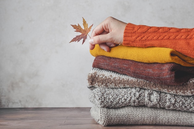 A females's hand holding a fallen leaf leaning on stack of knitted sweaters autumn color palette.