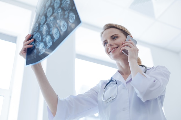 Feminine smart positive radiologist working at the x ray cabinet while examining brain x ray and using digital gadget for conversation