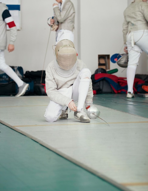 写真 レイピアと保護マスクが靴ひもを結ぶ白い衣装のフェンシング選手
