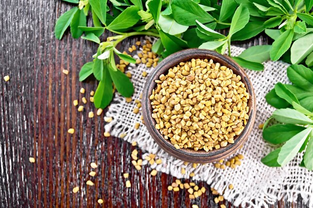 Fenugreek seeds in a bowl on burlap with green leaves on a wooden board from above