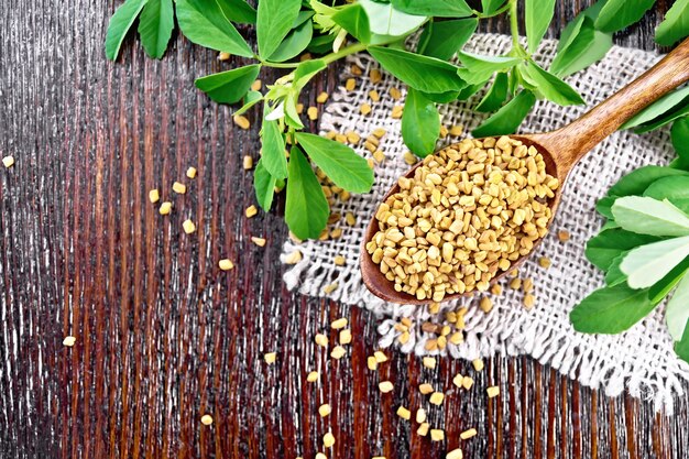 Photo fenugreek seeds in a spoon on a burlap napkin with green leaves on wooden board background from above