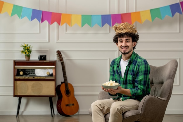 Festa junina ragazzo carino con cappello di paglia con radio retrò e bandiera colorata che tiene una ciotola di popcorn