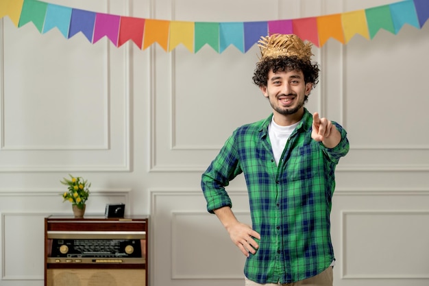 Festa junina cute young guy in straw hat with retro radio and colorful flags pointing finger