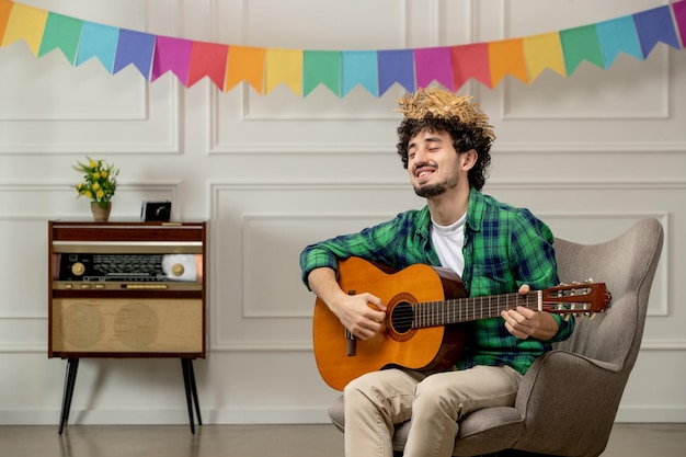 Festa junina schattige jonge kerel in strohoed met retro radio en kleurrijke vlaggen die op gitaar spelen