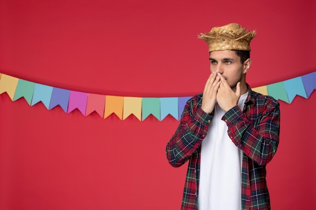 Foto festa junina sorridente ragazzo carino che indossa cappello di paglia festival brasiliano scioccato che copre la bocca