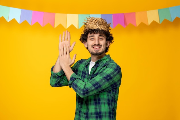 Festa junina молодой симпатичный парень в соломенной шляпе и разноцветных флагах бразильского фестиваля хлопает в ладоши