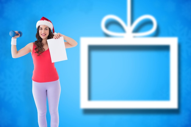 Photo festive athletic brunette holding page and dumbbell  against blurred christmas background