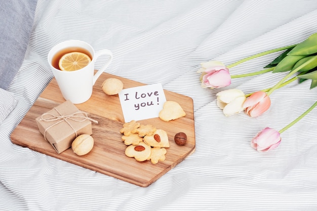 Festive breakfast in bed for Valentine's Day. Tea and cookies with your own hands in the form of hearts. A note on paper, a gift and flowers to your beloved girl.