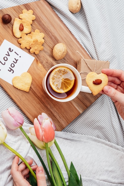 バレンタインデーのためのベッドでのお祝いの朝食。ハートの形で自分の手でお茶とクッキー。あなたの最愛の女の子への紙、贈り物、そして花へのメモ。