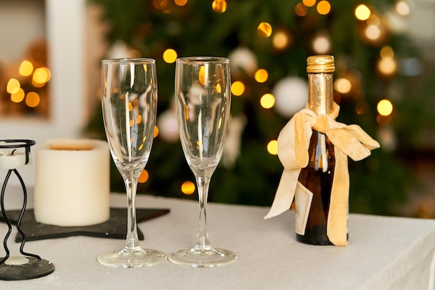 Festive Christmas background Cozy home evening Two glasses and a bottle of champagne on the background of a Christmas tree and bokeh of garlands