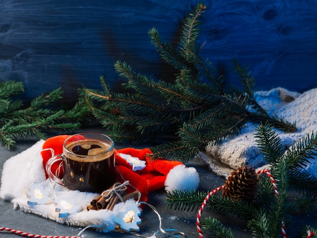 Festive christmas decor, a cup of hot espresso coffee on the table with christmas tree branches, santa's red hat, knitted clothes