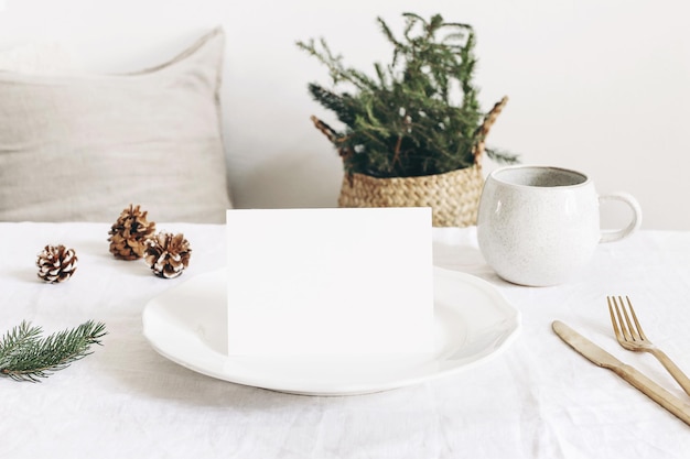 Festive Christmas winter table setting Golden cutlery spruce tree branch linen tablecloth porcelain dinner plate cup of coffee and silk pine cones on white table background Blank card mockup