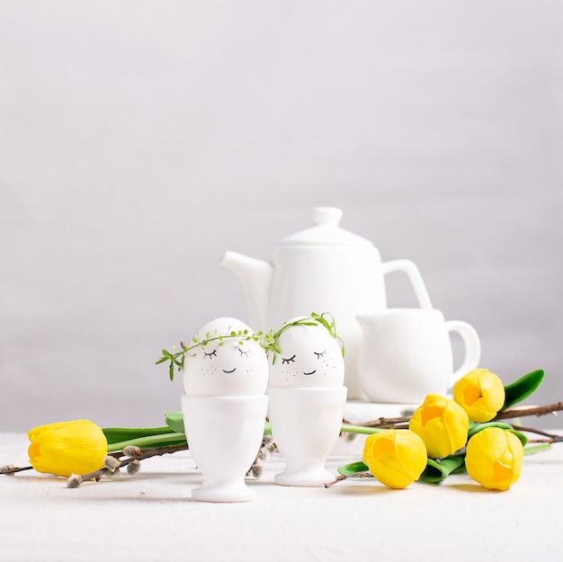 Праздничная композиция Пасхальные яйца в стаканах, белая посуда и желтые тюльпаны на белом столе Концепция счастливой Пасхи