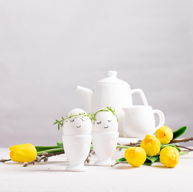 Festive composition Easter eggs in glasses white dishes and yellow tulips on a white table Happy Easter concept