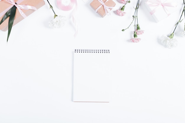 Праздничная композиция: блокнот, коробки с подарками, ленты и цветы лежат на белом столе