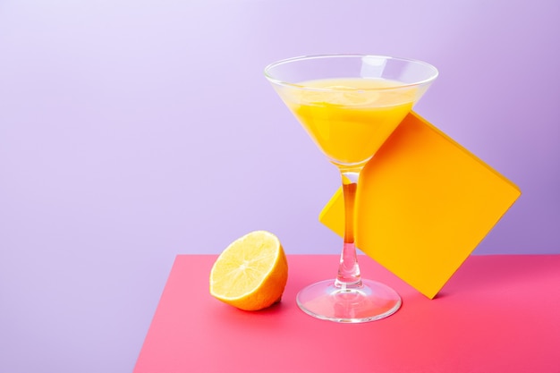 Праздничная композиция с коктейльным бокалом желтого напитка рядом с разрезанным пополам лимоном и желтой коробкой на ярком красочном фоне