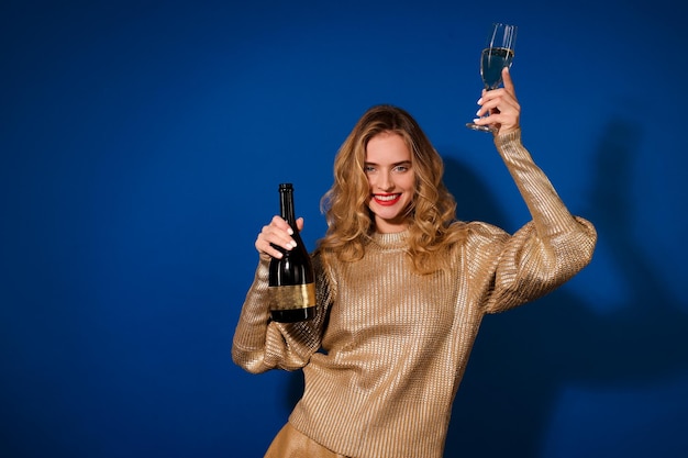 Festive drink. Shining long-haired girl with toothy smile showing bottle of champagne and raising glass looking at camera on blue background in studio