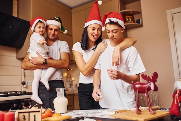 クリスマスの帽子をかぶったお祭りの家族は、キッチンで楽しんだり、食事を準備したりしています。