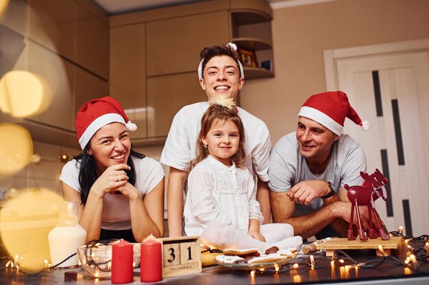 クリスマスの帽子をかぶったお祭りの家族は、キッチンで楽しんだり、食事を準備したりしています。