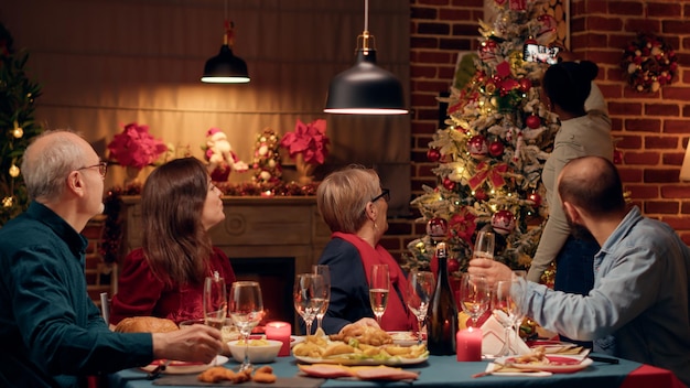 クリスマスの夕食のテーブルに座ってスマートフォンデバイスで自分撮り写真を撮るお祭りの家族。自分の写真を撮りながら冬のごちそうを祝う幸せな多様な人々。