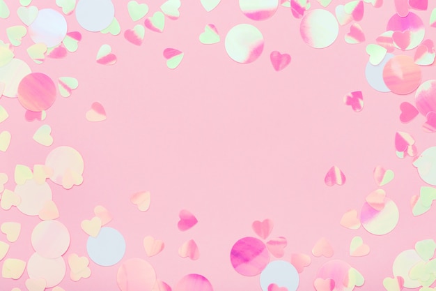 Festive frame of iridescent confetti sparkling on pink pastel background