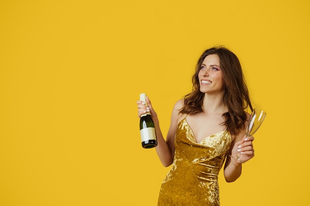 Festive mood happy lady in elegant evening dress with champagne bottle and glass in hand posing over